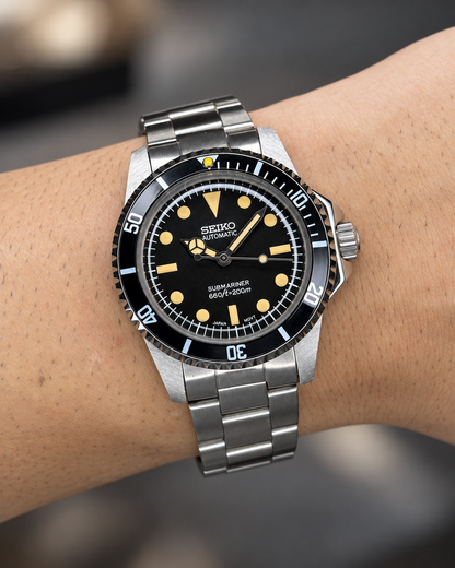 Submarino Vintage en la muñeca, con esfera negra, índices cálidos y brazalete de acero